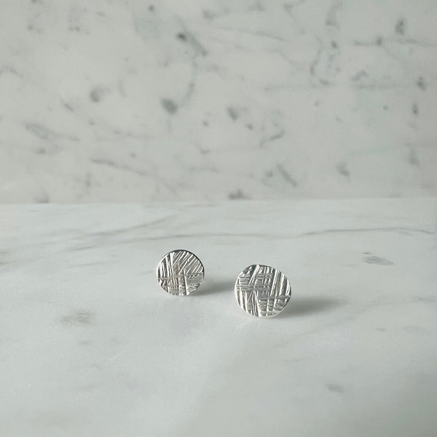A pair of small round silver stud earrings with a textured, hammered surface, resting on a white marble surface against a matching marble background.