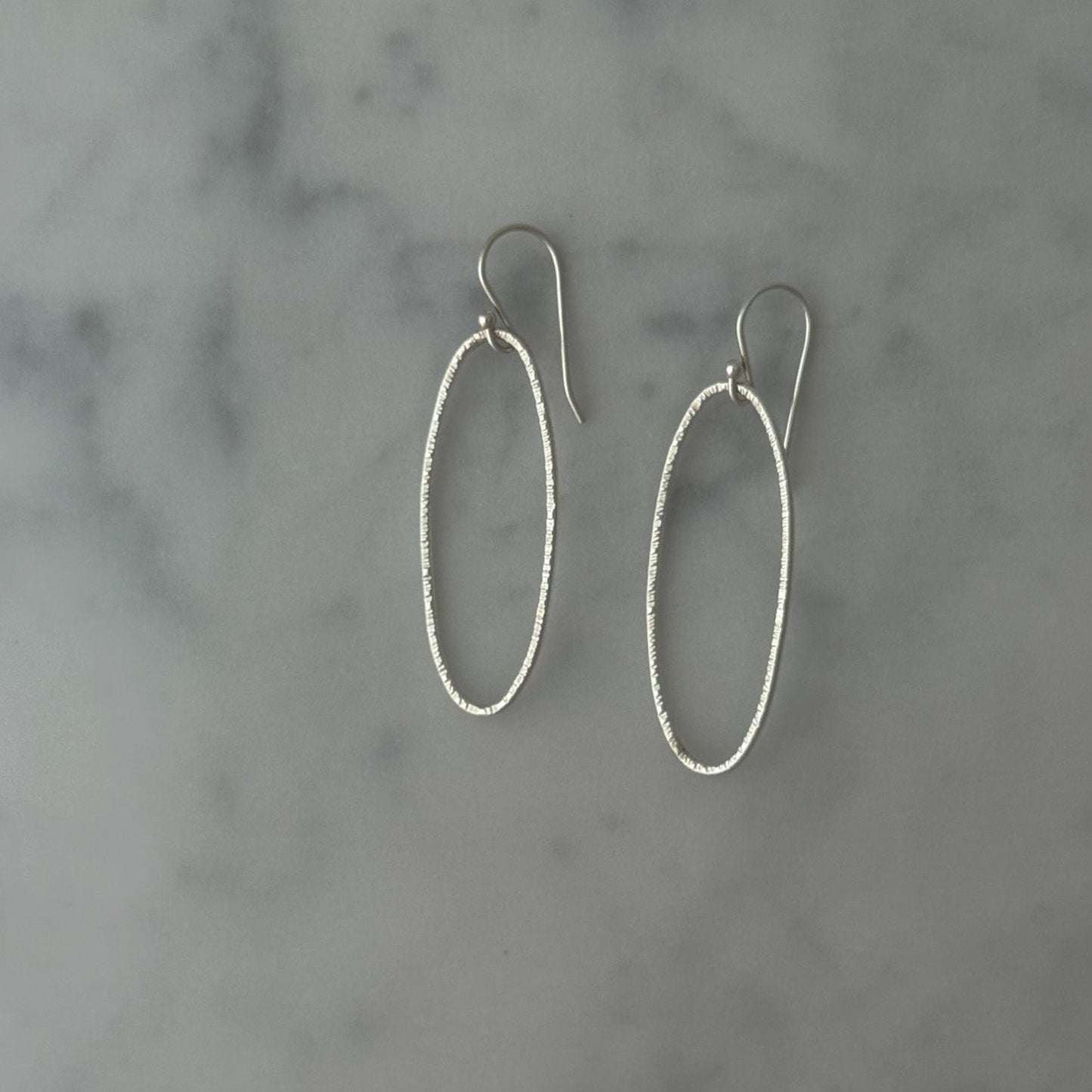 A pair of silver oval drop earrings with textured wire-like edges and fishhook backings, displayed on a light gray and white marble surface.