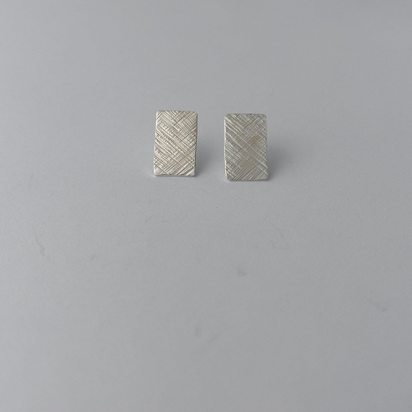 A pair of small rectangular silver earrings with a textured, woven-look surface, displayed on a plain light gray background.