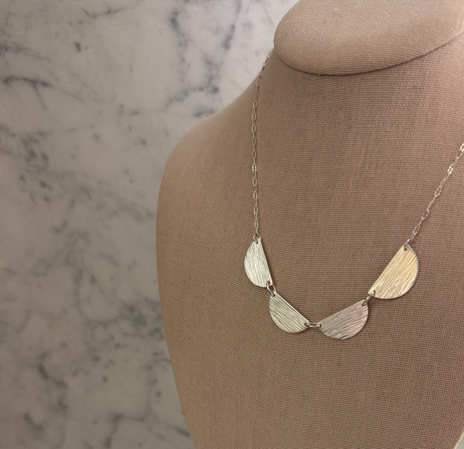 A silver-toned necklace with five textured, fan-shaped metal discs on a chain, displayed on a beige fabric jewelry bust against a light marble-patterned background.