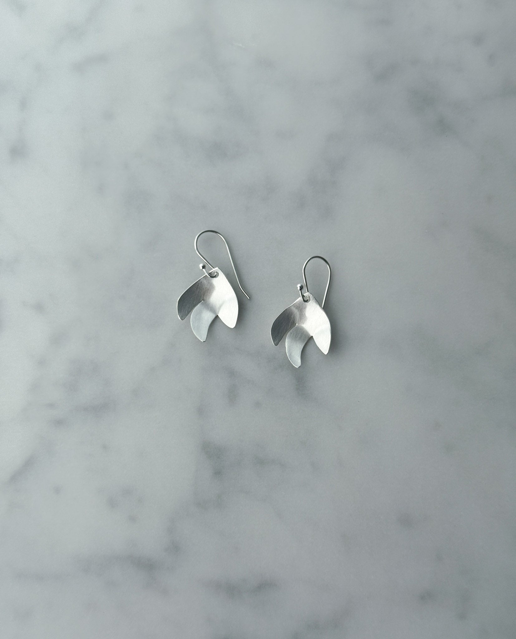 A pair of silver dangle earrings with a polished, leaf-like organic shape, resting on a light gray and white marble surface.