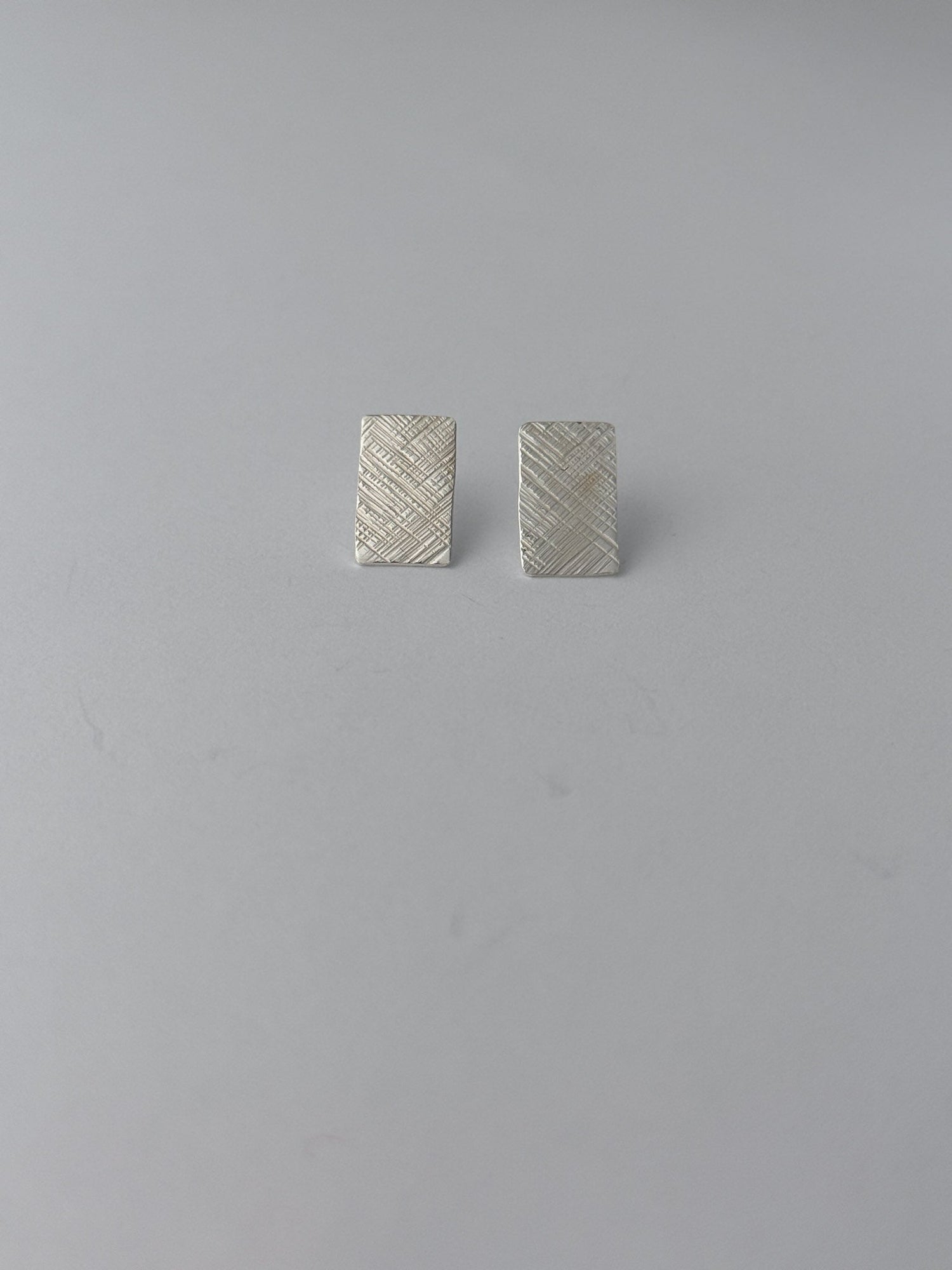 A pair of small rectangular silver earrings with a textured, woven-look surface, displayed on a plain light gray background.