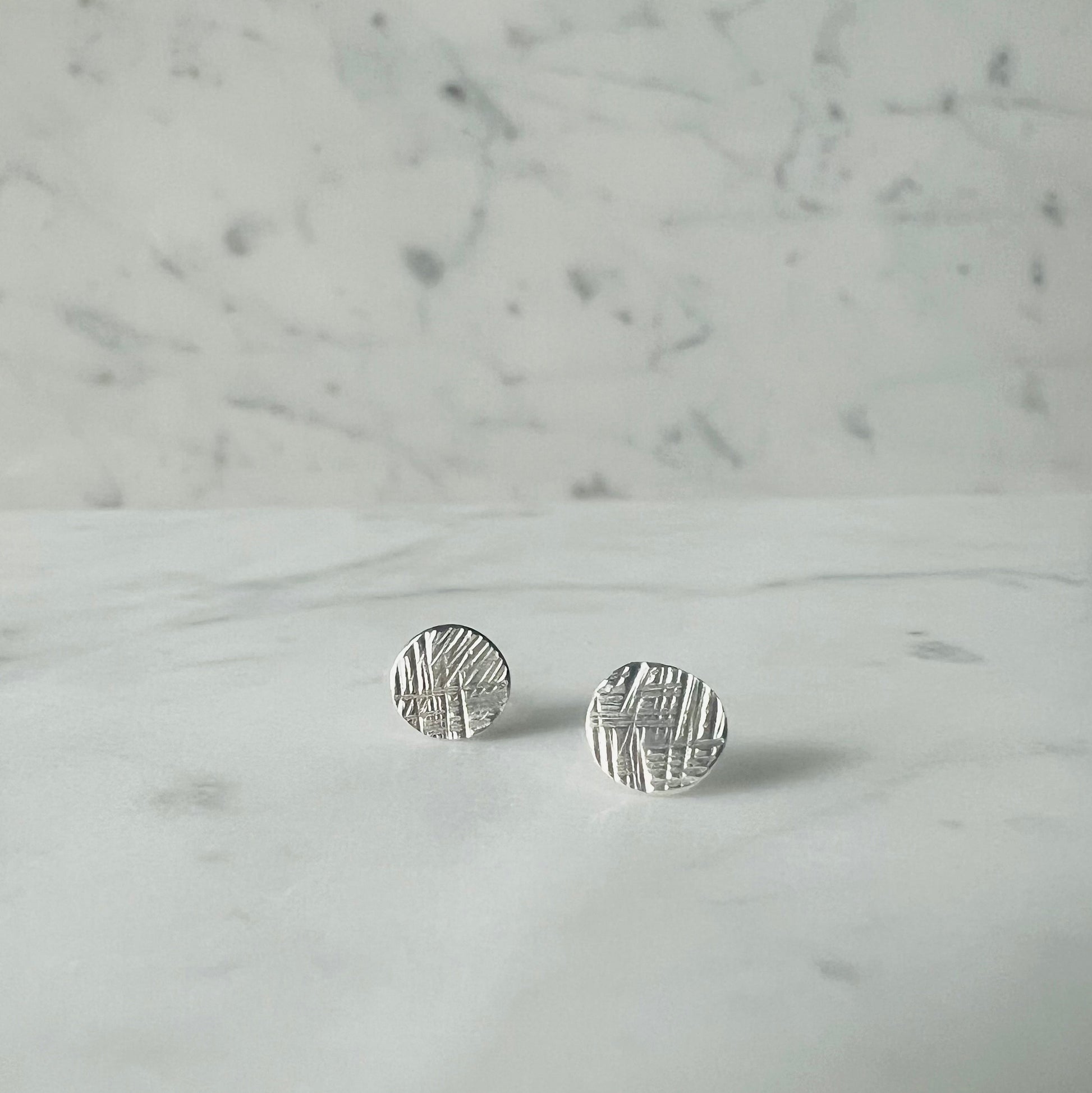 A pair of small round silver stud earrings with a textured, hammered surface, resting on a white marble surface against a matching marble background.