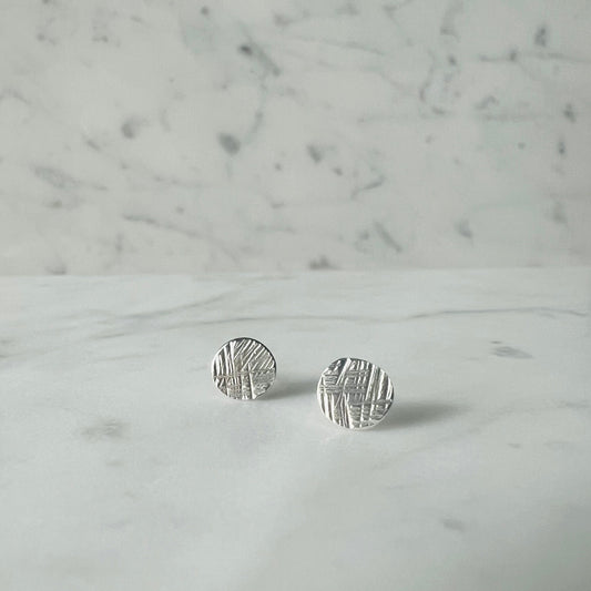 A pair of small round silver stud earrings with a textured, hammered surface, resting on a white marble surface against a matching marble background.