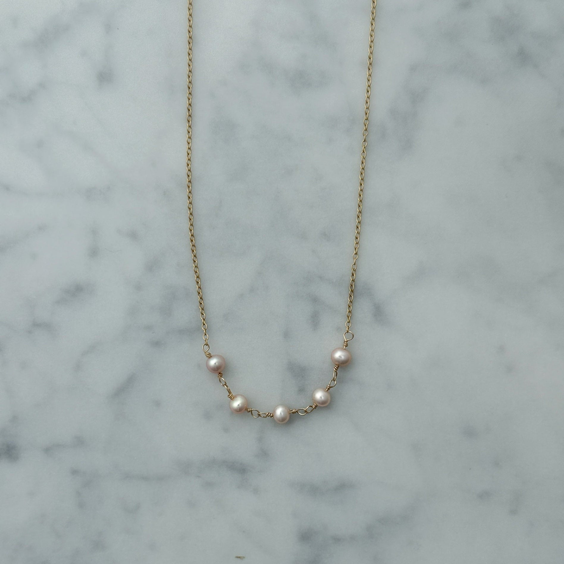 A delicate gold chain necklace featuring five small, lustrous white pearls spaced along the chain, displayed on a white and gray marble surface.