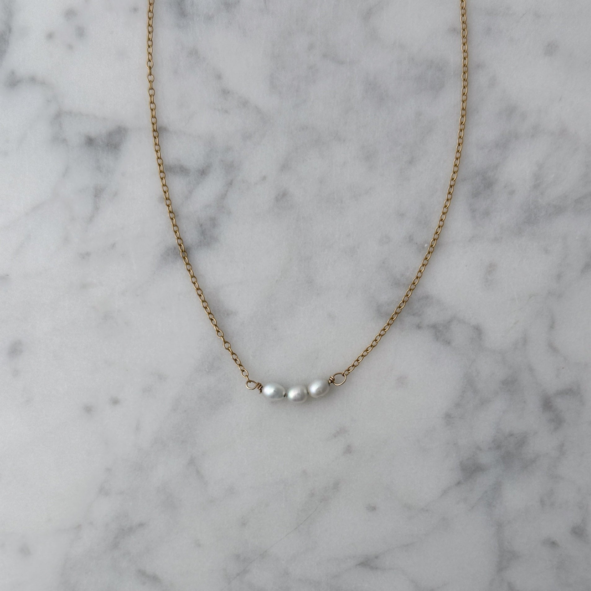 A delicate gold chain necklace with three small white pearls centered on a white and gray marble surface.
