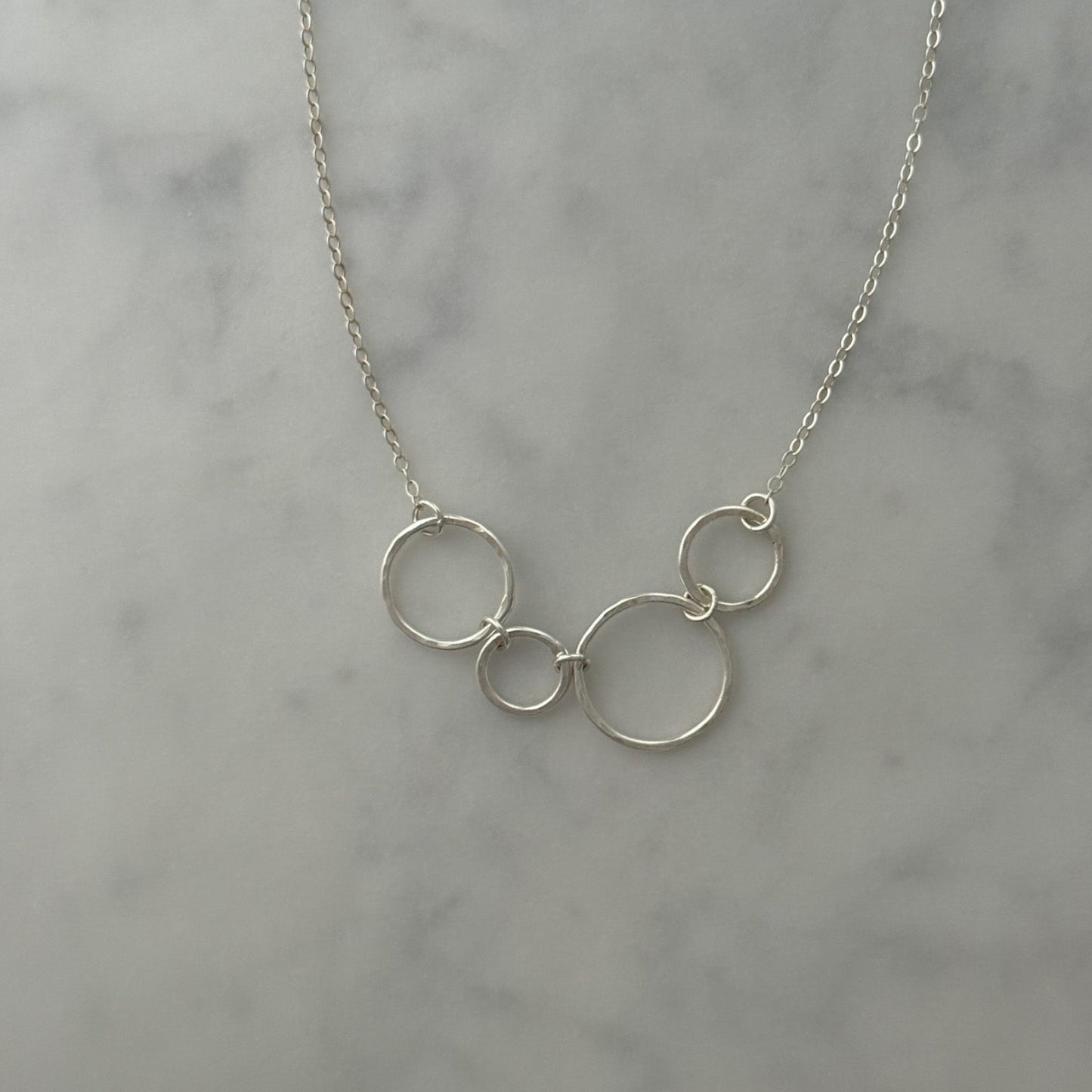 A delicate silver chain necklace featuring three interlocking open circle pendants in varying sizes, displayed on a light gray marble surface.