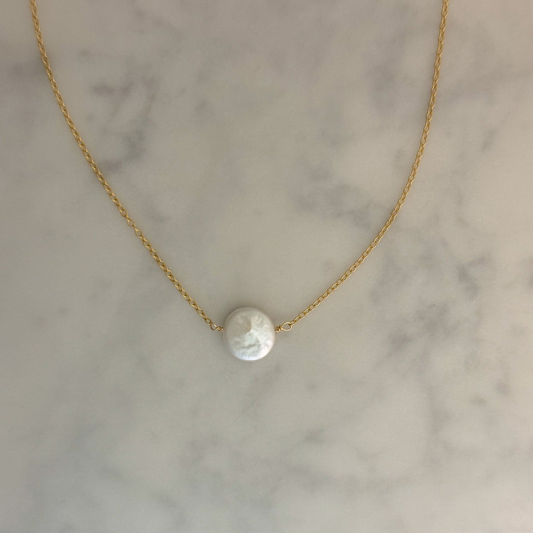 A delicate gold chain necklace with a single round white pearl pendant, displayed on a light gray and white marble surface.