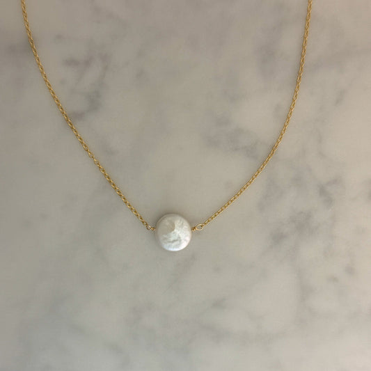 A delicate gold chain necklace with a single round white pearl pendant, displayed on a light gray and white marble surface.
