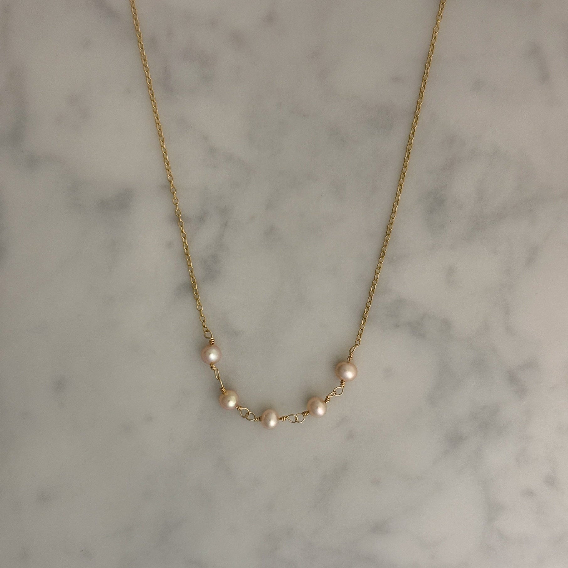 A delicate gold chain necklace featuring five small, pale pinkish-white pearls spaced along the chain, displayed on a light gray and white marble surface.