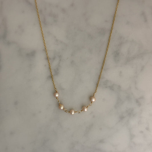 A delicate gold chain necklace featuring five small, pale pinkish-white pearls spaced along the chain, displayed on a light gray and white marble surface.