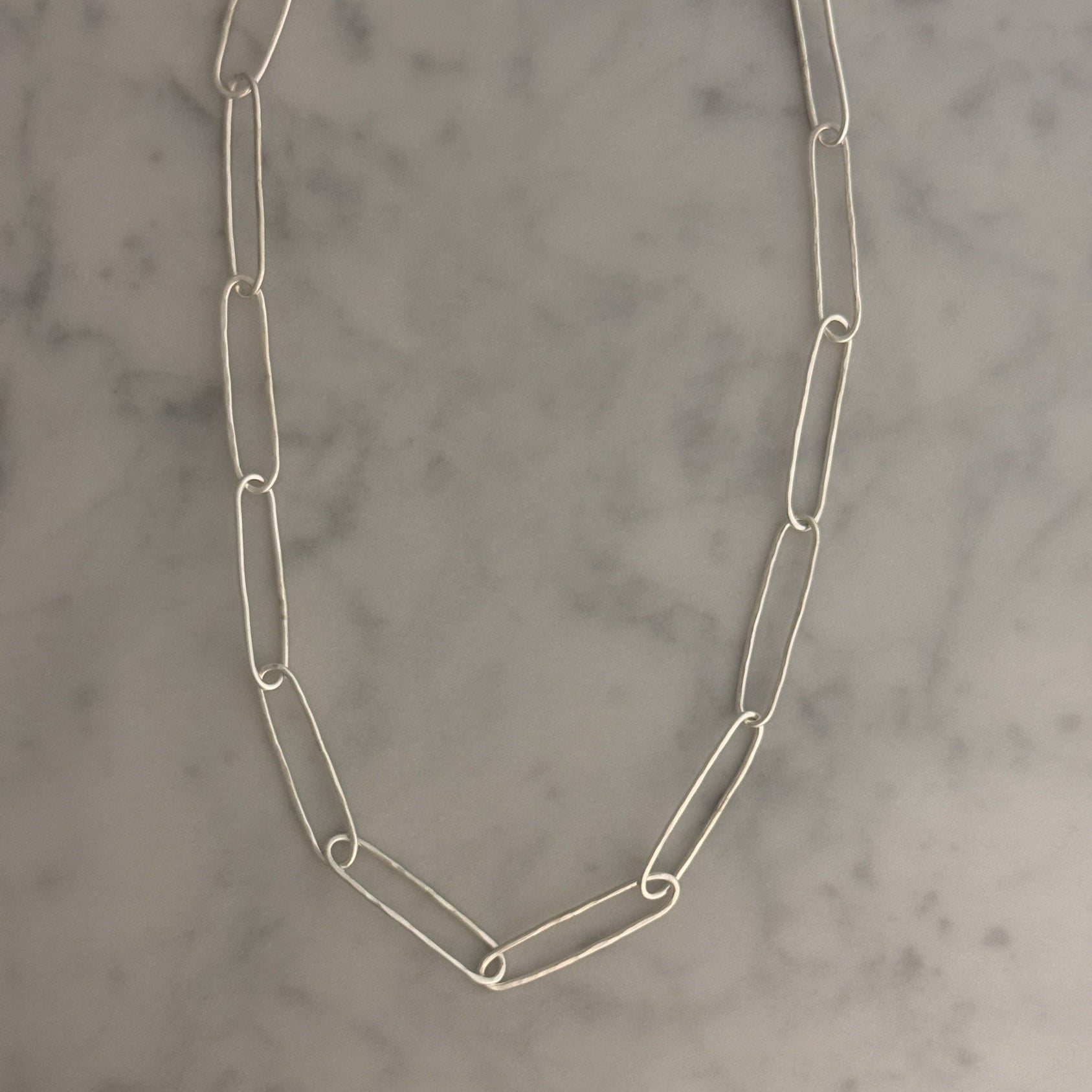 A silver paperclip chain necklace laid flat on a light gray and white marble surface.