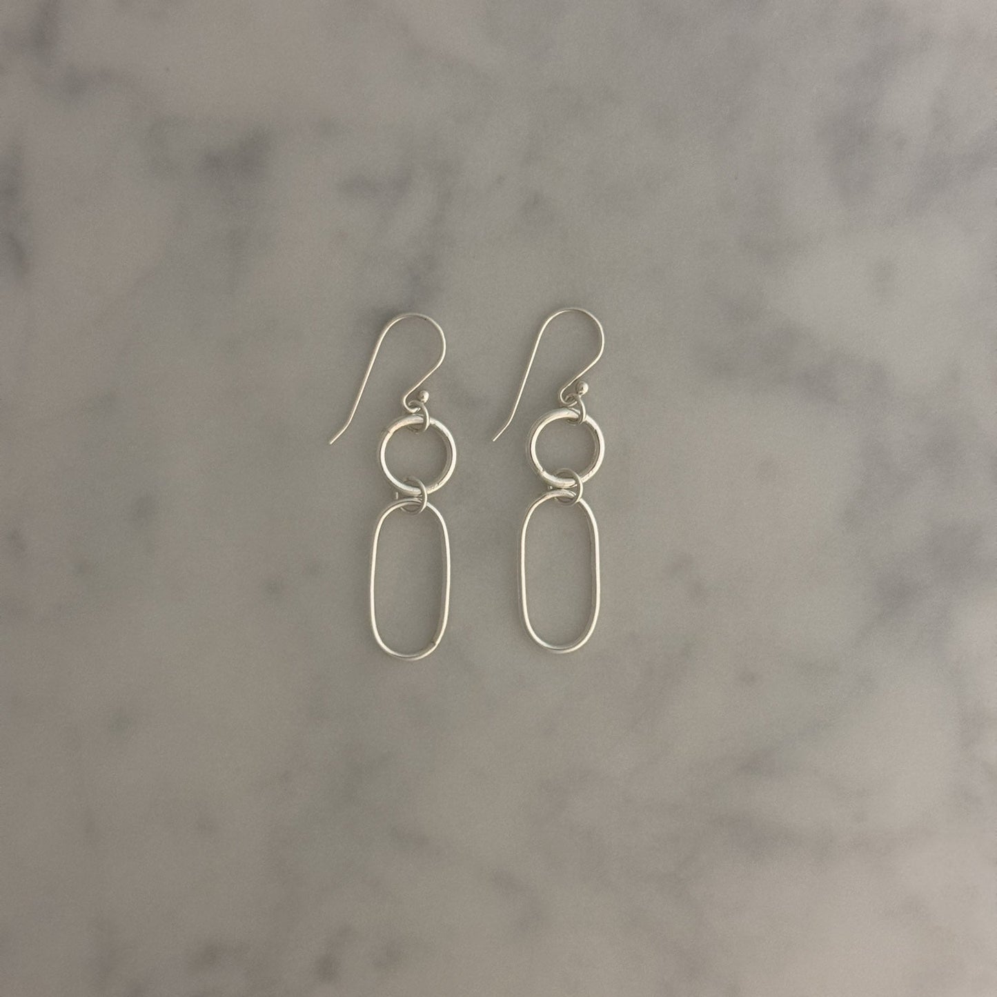 A pair of minimalist silver drop earrings with circular and elongated oval wireframe details, resting on a light gray and white marble surface.