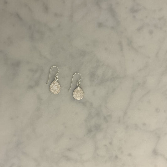 A pair of silver teardrop dangle earrings with a textured hammered finish, resting on a light gray and white marble surface.