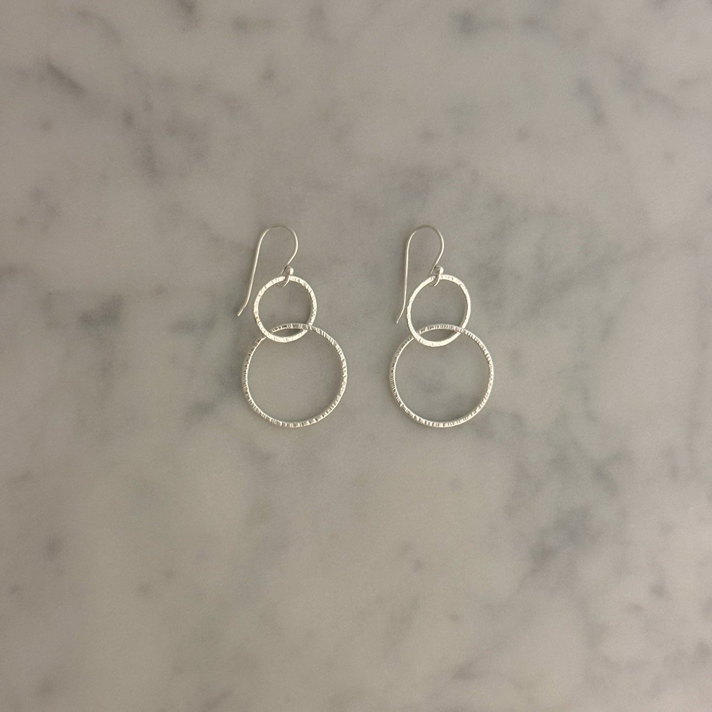 A pair of silver dangle earrings with interlocking textured hoop designs, displayed on a light gray and white marble surface.