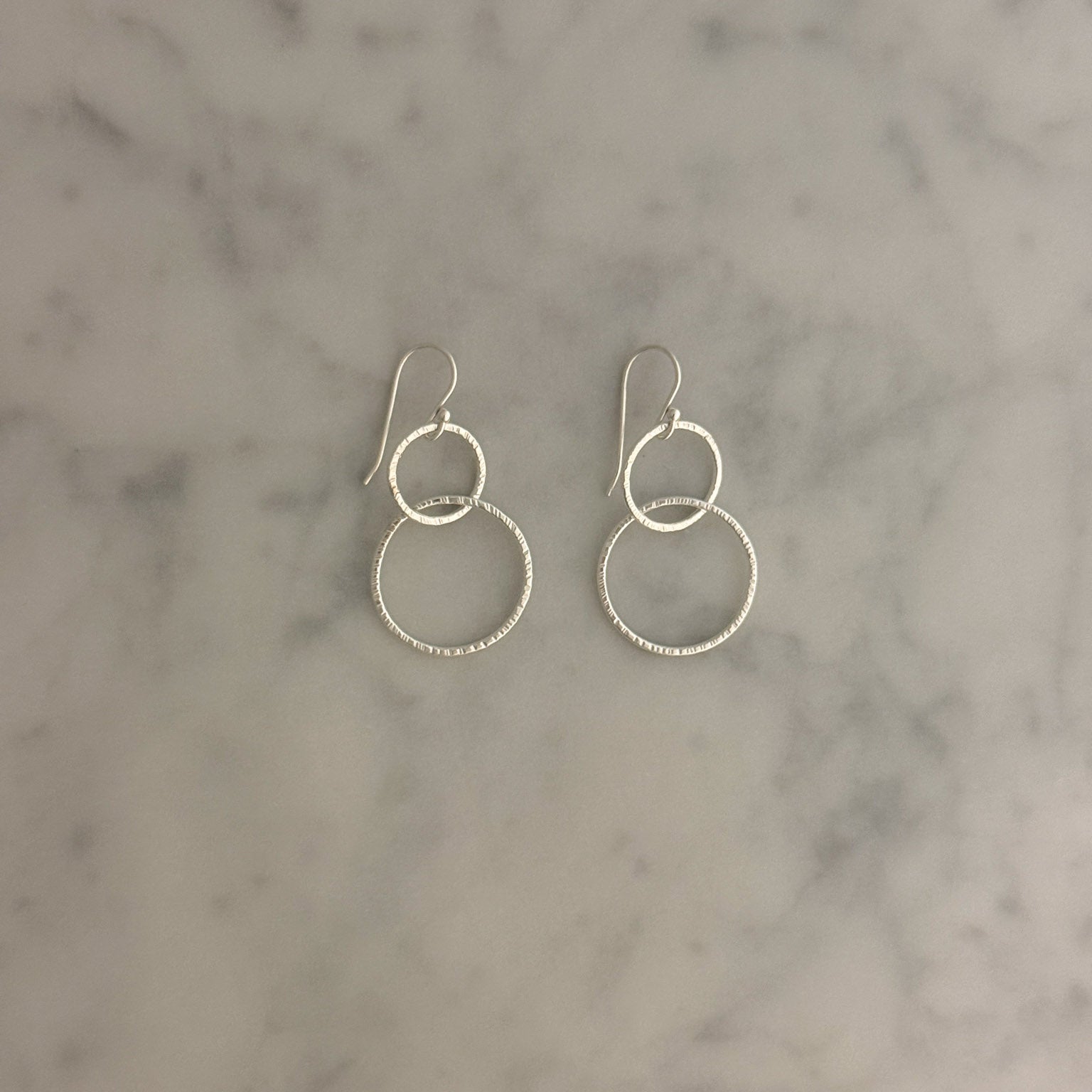 A pair of silver dangle earrings with interlocking textured hoop designs, displayed on a light gray and white marble surface.
