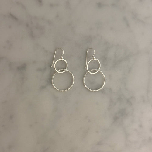A pair of silver dangle earrings with interlocking textured hoop designs, displayed on a light gray and white marble surface.