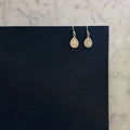 A pair of silver teardrop dangle earrings with textured, glittery gold-toned drops, displayed on a black card over a white marble surface.