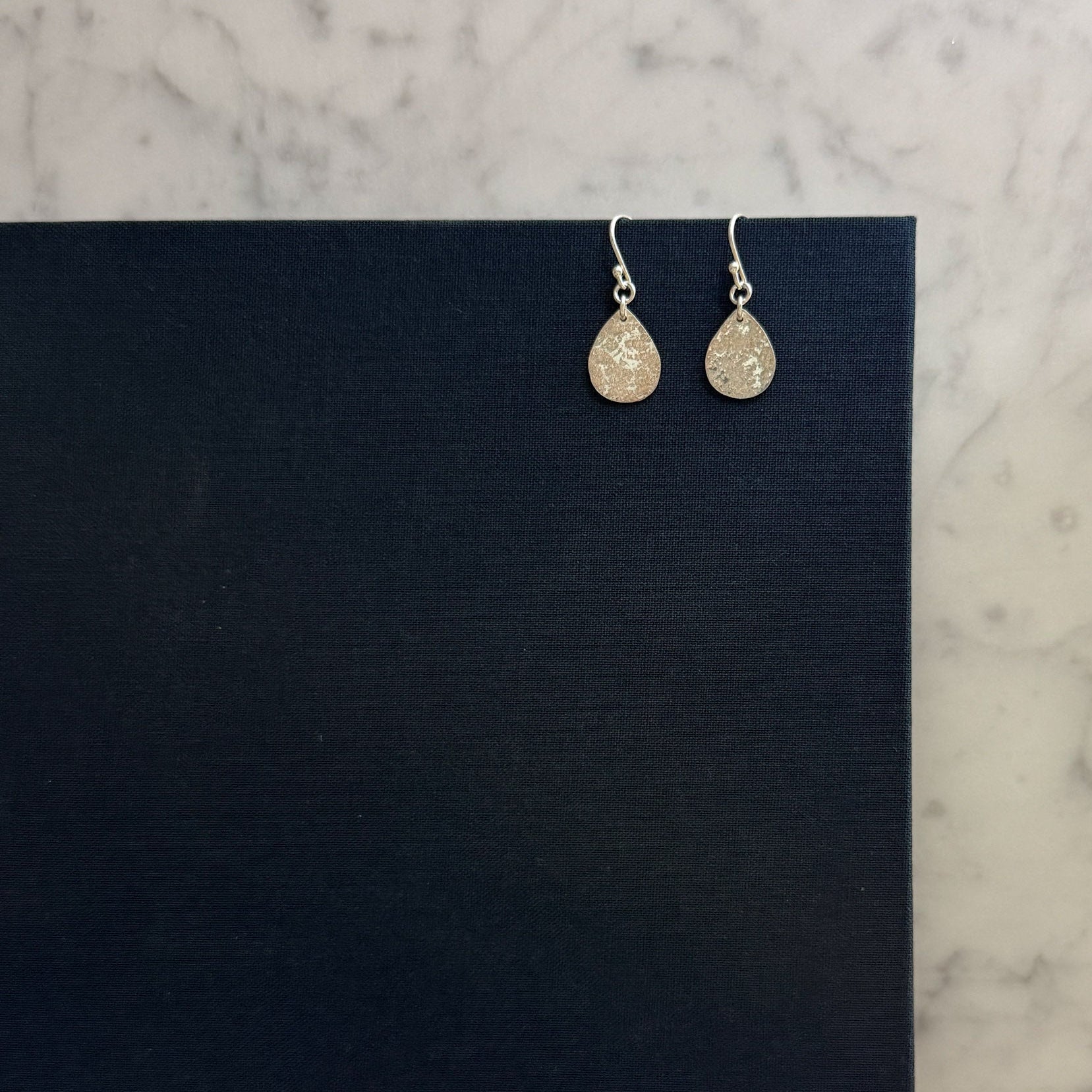 A pair of silver teardrop dangle earrings with textured, glittery gold-toned drops, displayed on a black card over a white marble surface.