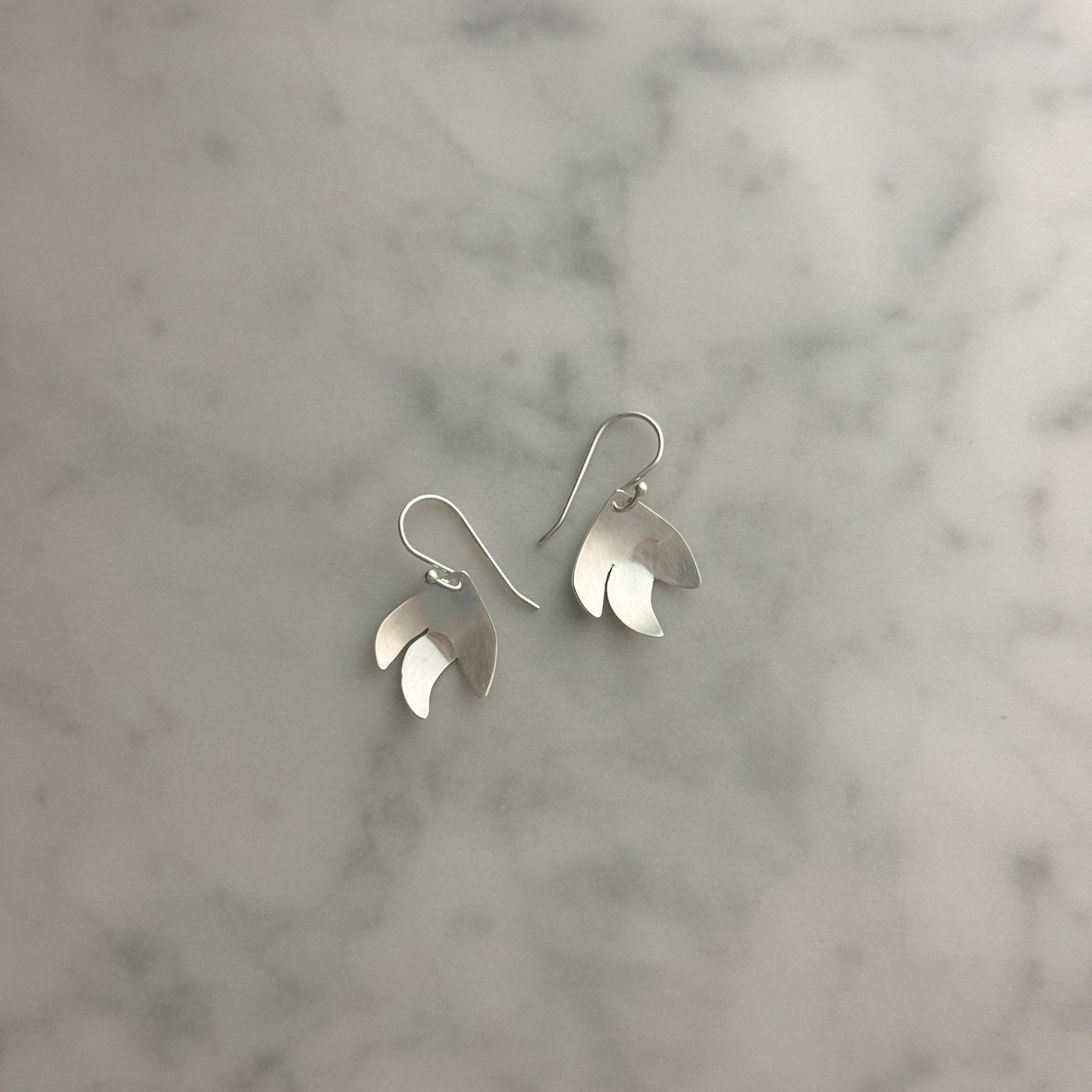 A pair of silver dangle earrings with a layered, petal-like design, displayed on a light gray and white marble surface.