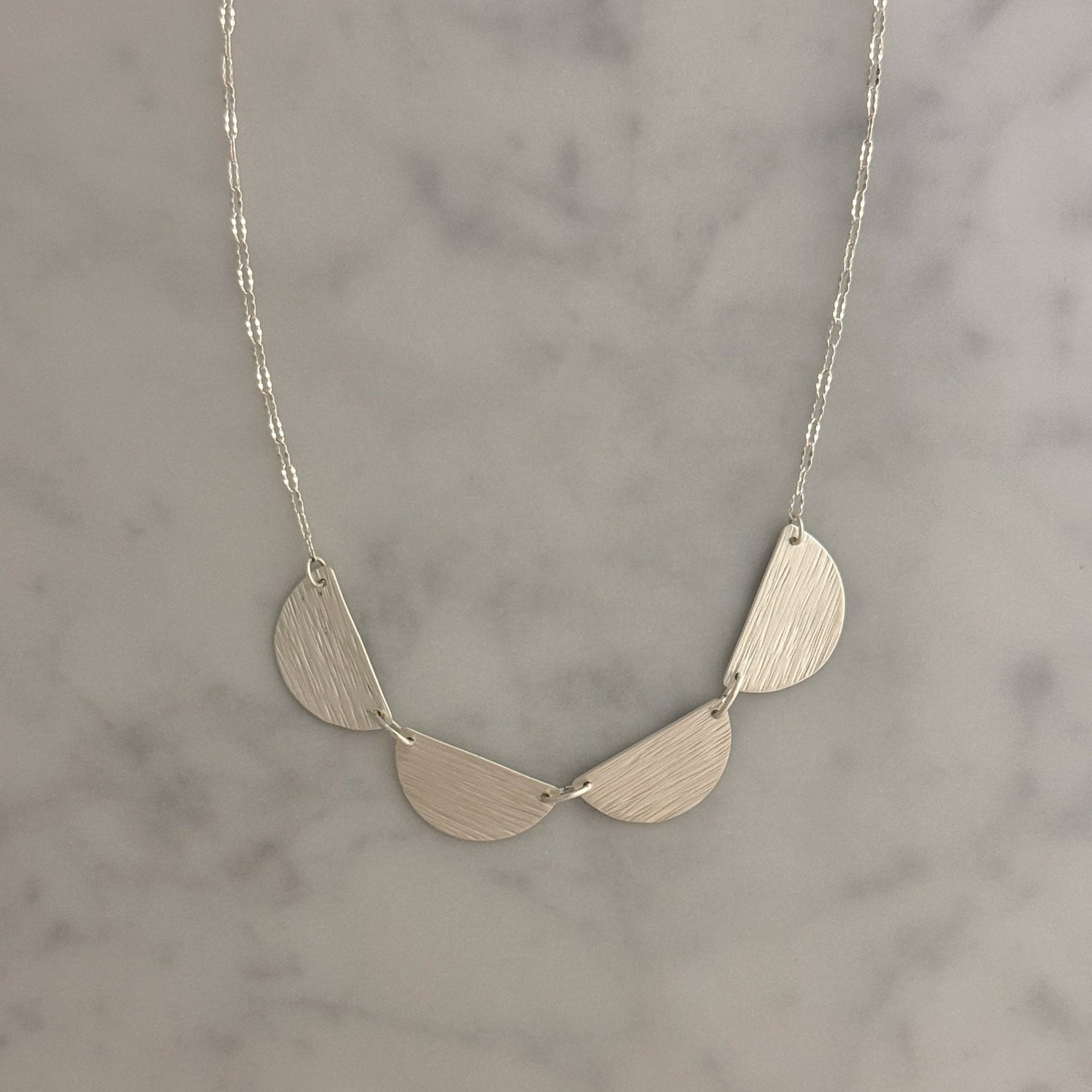 A silver necklace with three textured, semi-circular leaf-like drops hanging from a delicate silver chain, displayed on a light gray marble surface.