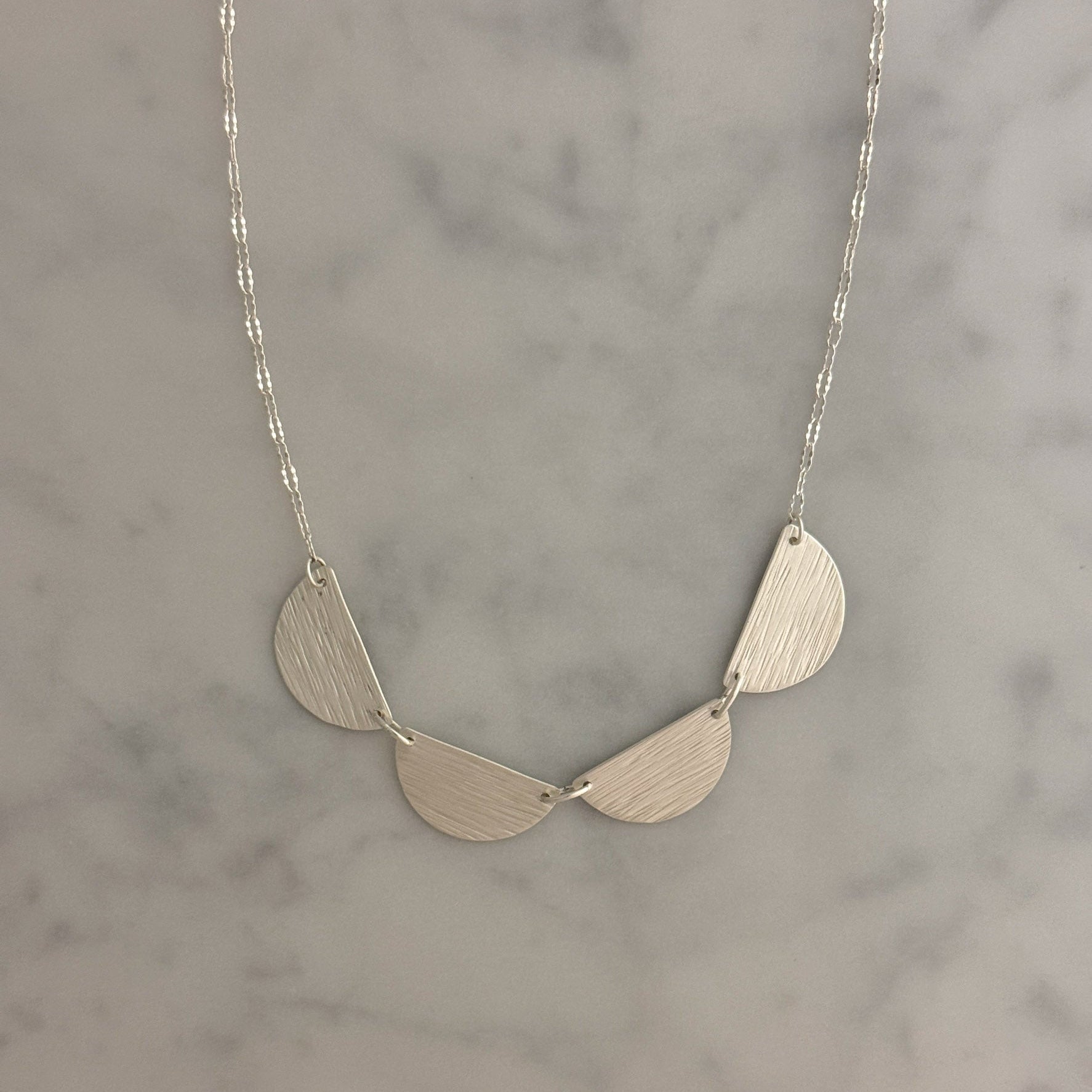 A silver necklace with three textured, semi-circular leaf-like drops hanging from a delicate silver chain, displayed on a light gray marble surface.