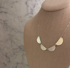 A silver-toned necklace with five textured, fan-shaped metal discs on a chain, displayed on a beige fabric jewelry bust against a light marble-patterned background.