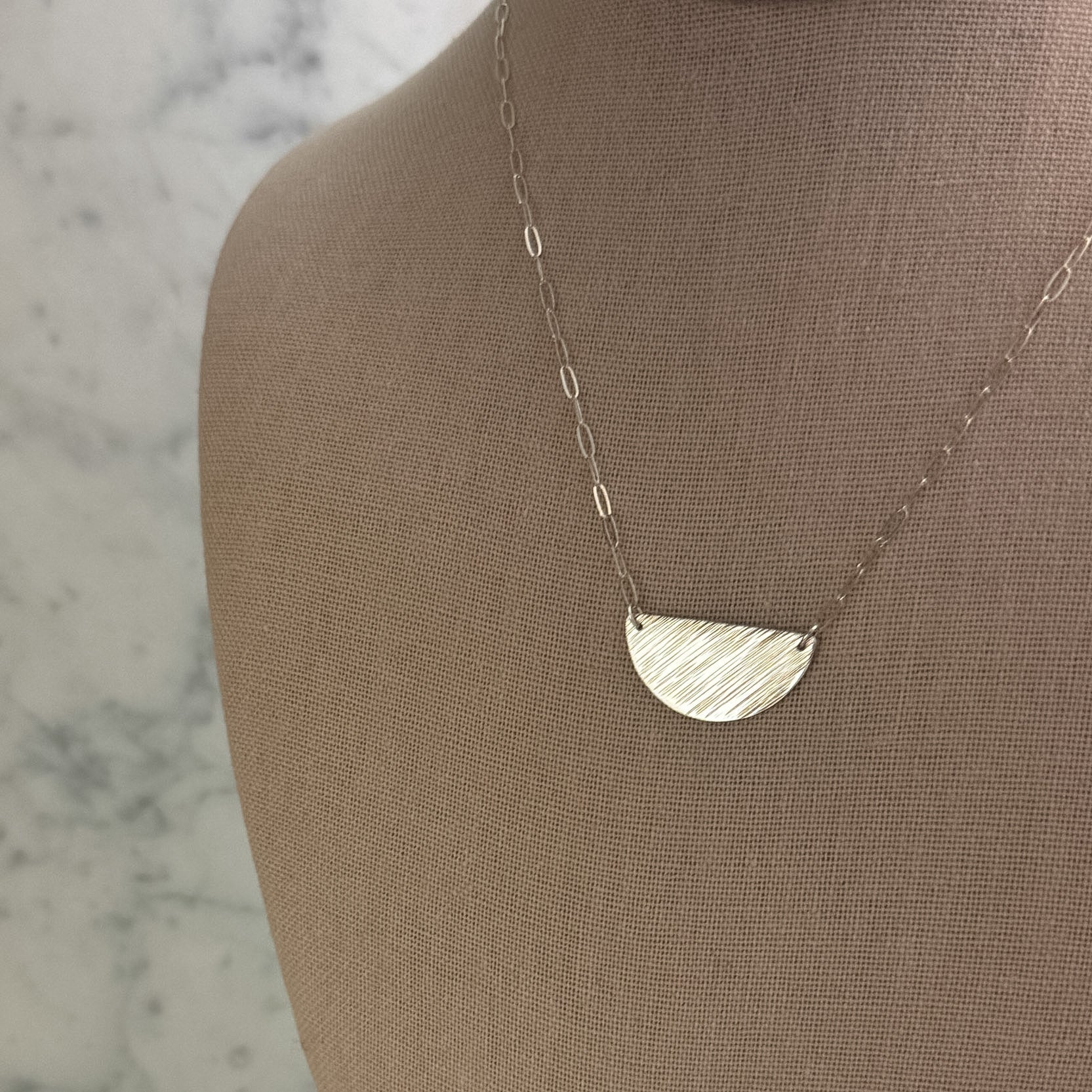 A silver necklace with a textured, semi-circular half-moon pendant displayed on a beige fabric jewelry bust against a light marble background.