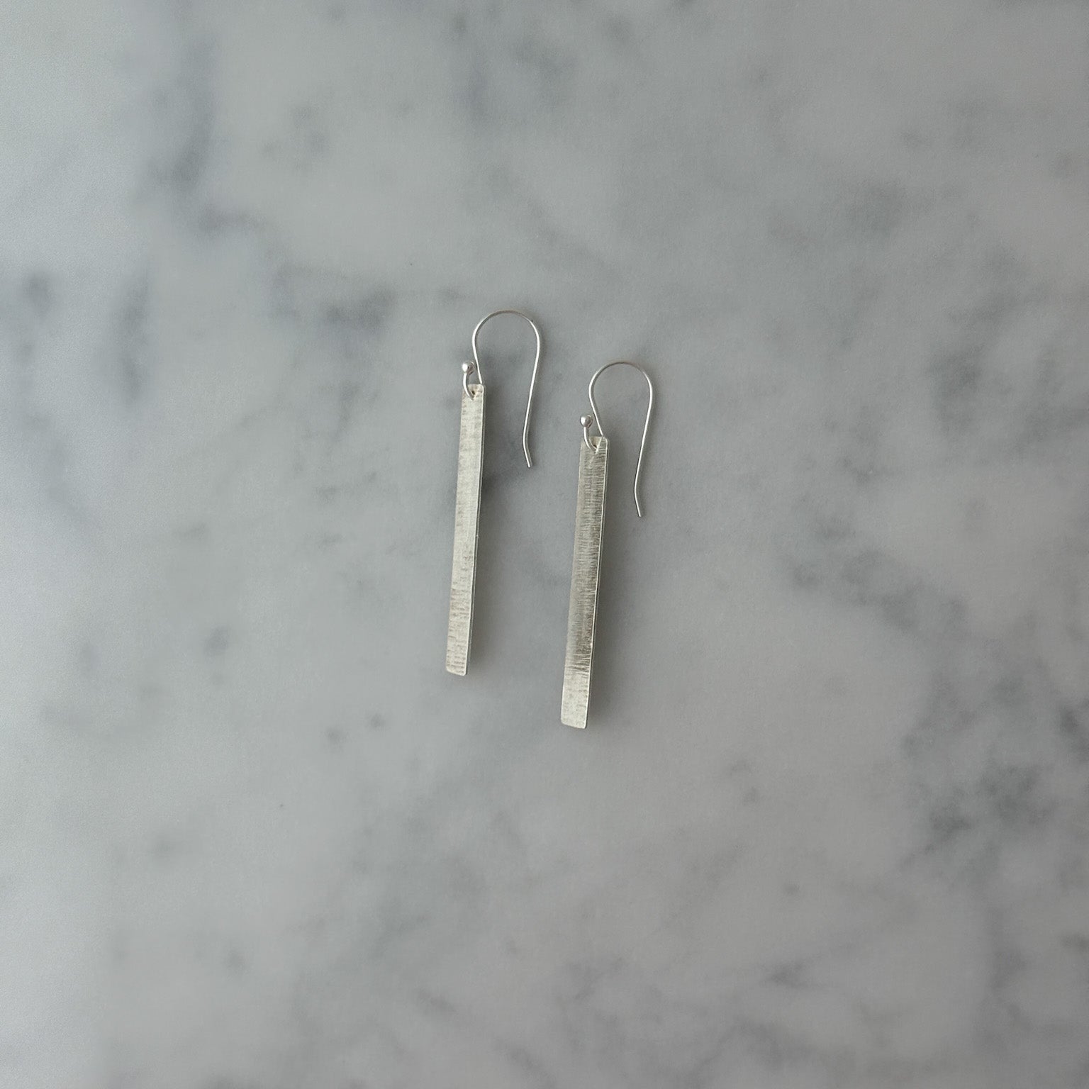 A pair of silver drop earrings with textured rectangular bars and fishhook backs, displayed on a white and gray marble surface.