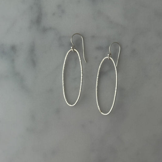 A pair of silver oval drop earrings with textured wire-like edges and fishhook backings, displayed on a light gray and white marble surface.