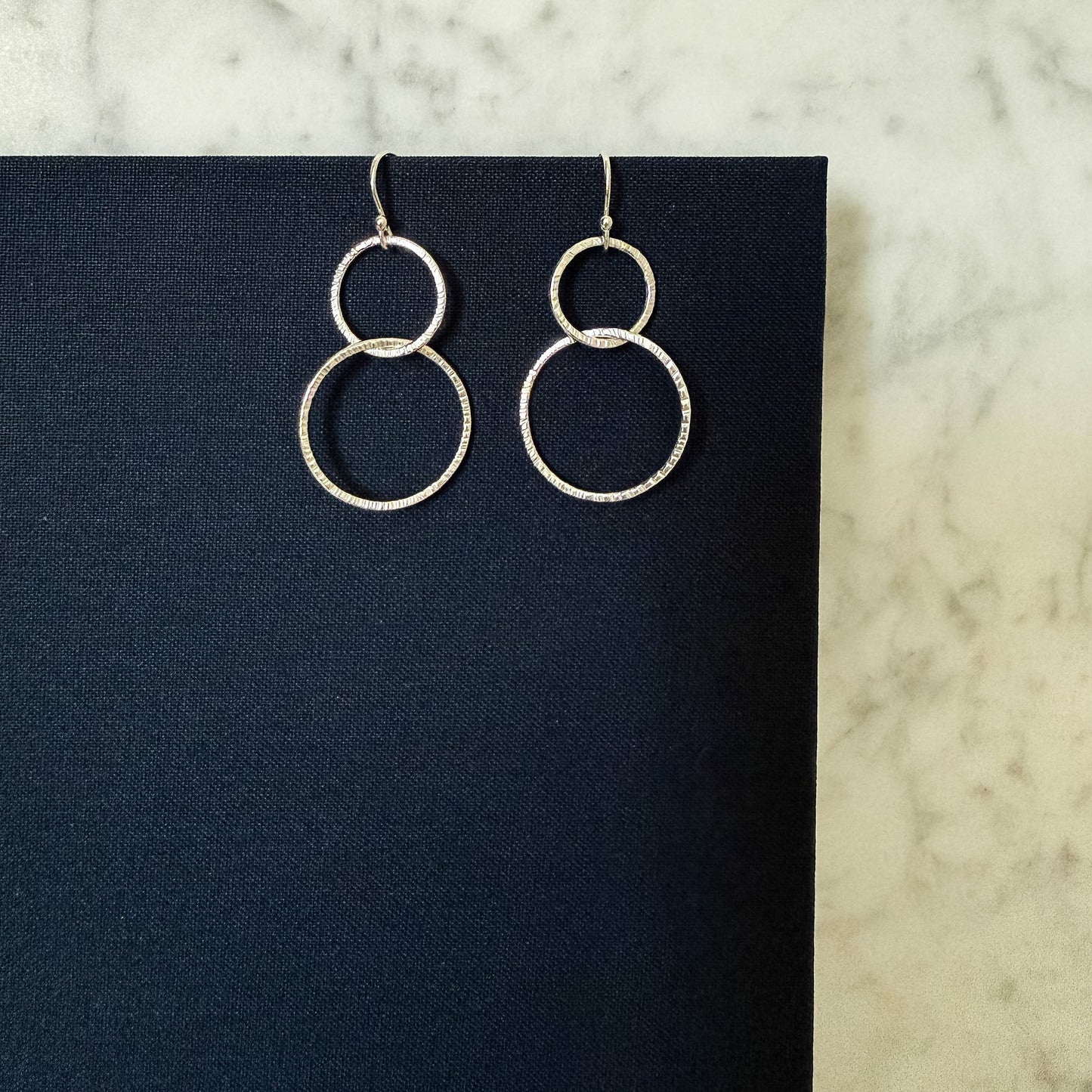 A pair of silver-toned double-ring dangle earrings with a textured, pave-like finish, displayed on a black rectangular card against a light marble background.