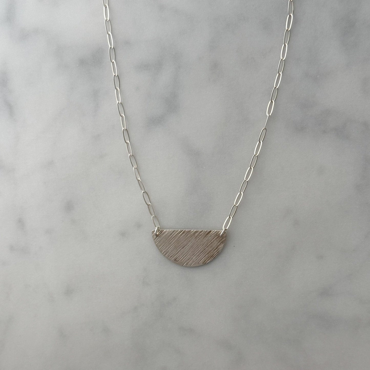 A silver necklace with a textured, semi-circular half-moon pendant on a delicate paperclip chain, displayed on a light gray and white marble surface.