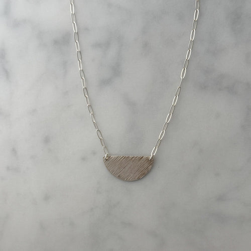 A silver necklace with a textured, semi-circular half-moon pendant on a delicate paperclip chain, displayed on a light gray and white marble surface.