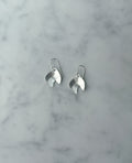 A pair of silver dangle earrings with a polished, leaf-like organic shape, resting on a light gray and white marble surface.