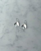 A pair of silver dangle earrings with a polished, leaf-like organic shape, resting on a light gray and white marble surface.