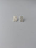 A pair of small rectangular silver earrings with a textured, woven-look surface, displayed on a plain light gray background.