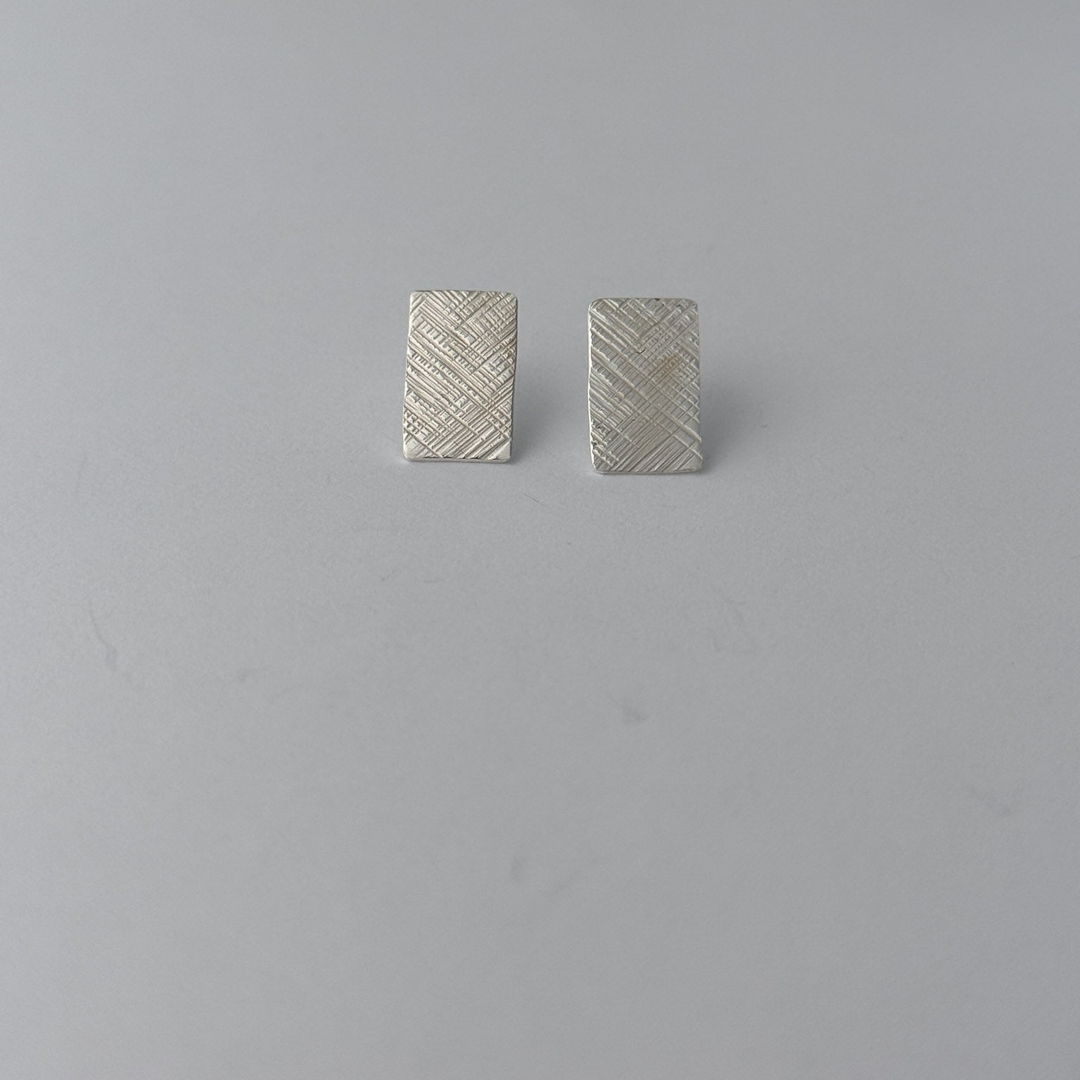 A pair of small rectangular silver earrings with a textured, woven-look surface, displayed on a plain light gray background.