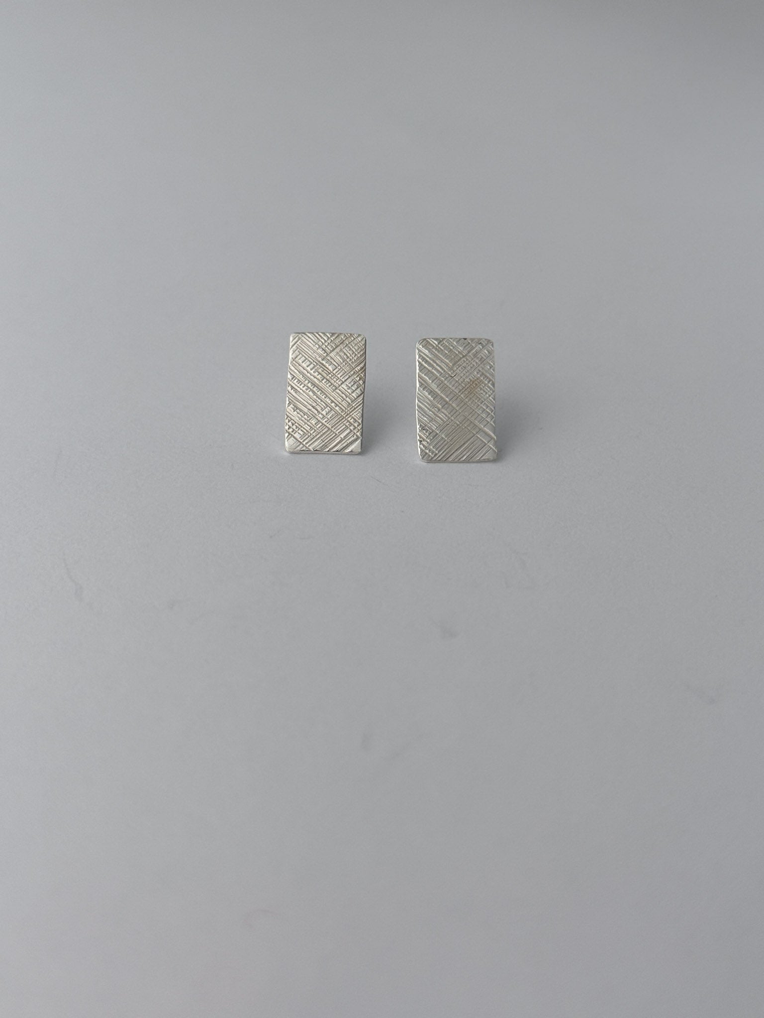 A pair of small rectangular silver earrings with a textured, woven-look surface, displayed on a plain light gray background.