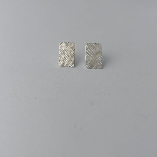 A pair of small rectangular silver earrings with a textured, woven-look surface, displayed on a plain light gray background.